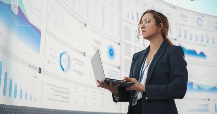 Successful Hispanic 业务woman Holding Laptop Computer And Reviewing Financial Reports On Digital Screen In Monitoring Office. 人工智能公司深度学习的女性领导者.